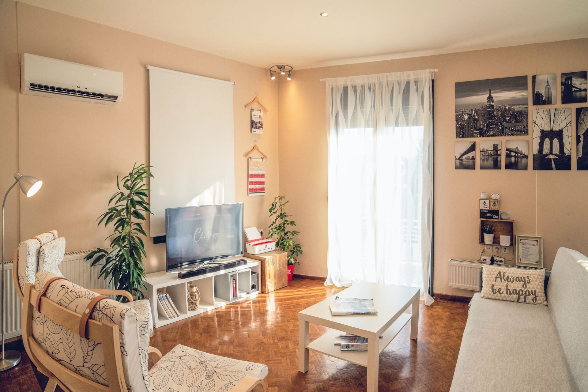 Cozy living room with light furniture, TV, plants, and sheer curtains letting in sunlight.