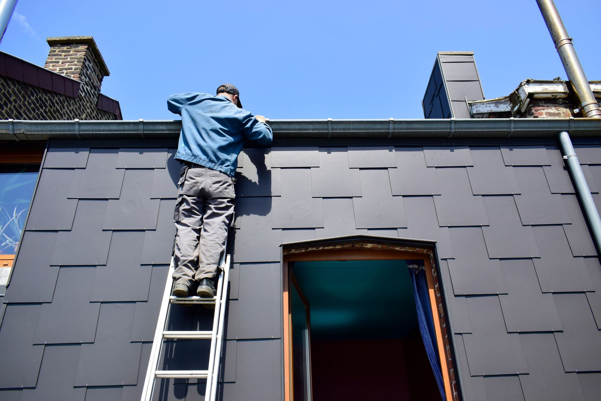 A caucasian man standing on the aluminium ladder and cleaning a roof gutters in sunshine day for preparation for water runoff in raining season.